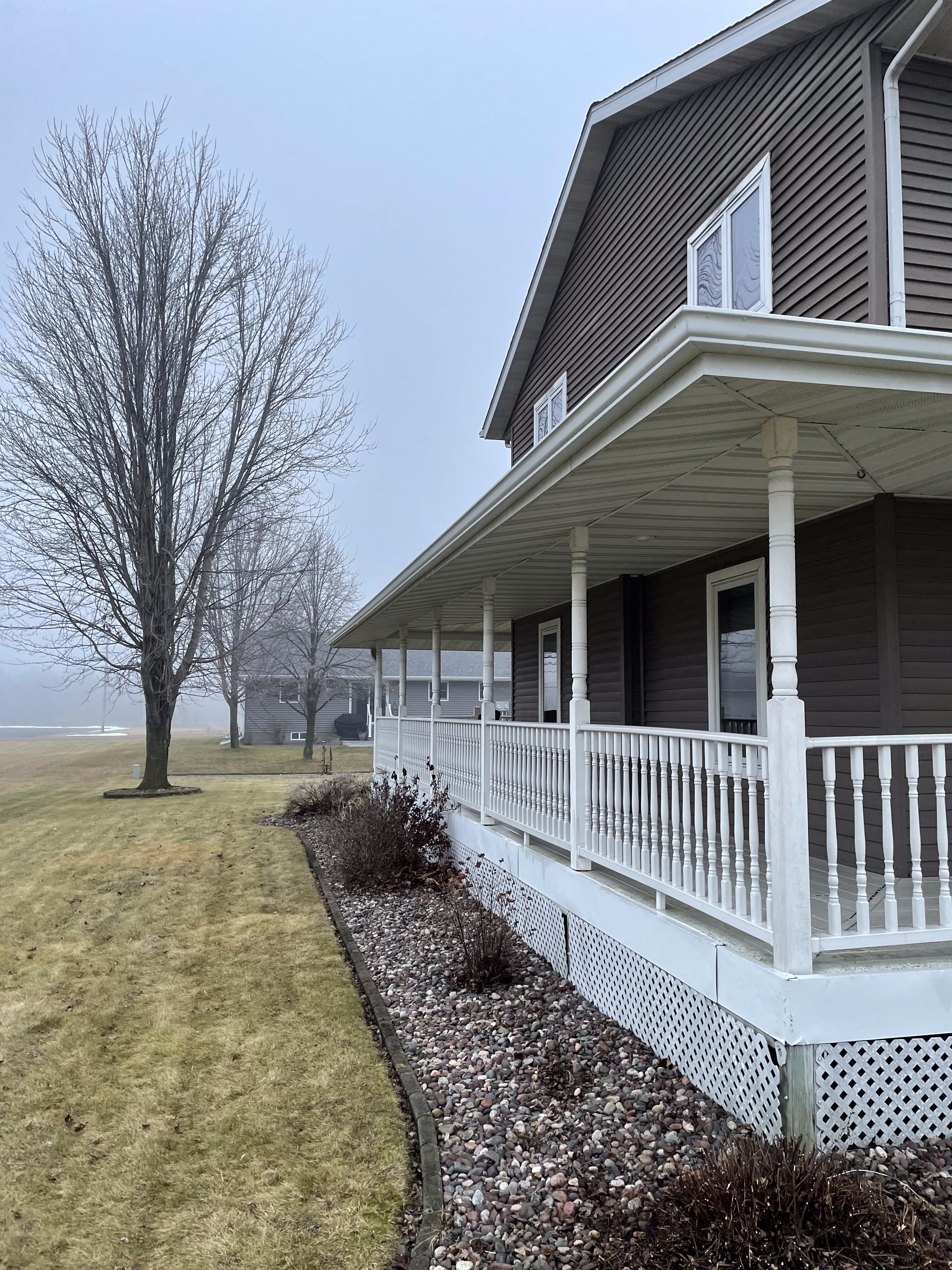 Two-story house with brown siding, white soffit and fascia installed along the roofline and porch ceiling, white porch railing with decorative posts, landscaped yard with bare trees and rock bed, cloudy sky over a residential area in Wisconsin.
